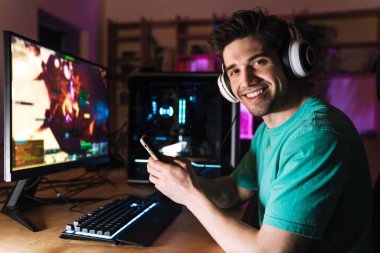 Evde bilgisayar oyunu oynarken kulaklıkla cep telefonu kullanan neşeli genç bir adam görüntüsü