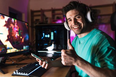 Neşeli bir adamın evde bilgisayar oyunu oynarken baş parmağını göstermesi ve cep telefonu kullanması