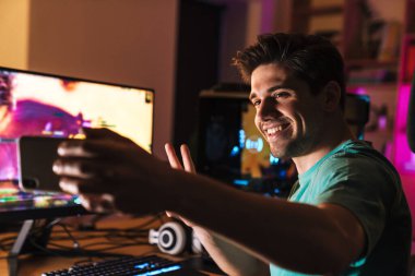 Neşeli bir adamın evde video oyunu oynarken cep telefonuyla fotoğraf çektiği ve barış işareti yaptığı görüntüsü