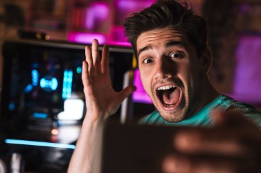 Memnuniyet verici bir adamın evde video oyunu oynarken cep telefonuyla çığlık atıp selfie çekerken görüntüsü