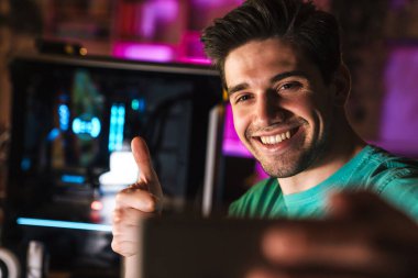 Neşeli bir adamın evde video oyunu oynarken baş parmağını gösterip cep telefonuyla selfie çekerken görüntüsü