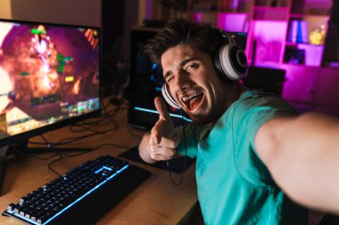 Evdeki bilgisayarda bilgisayar oyunu oynarken kamerayı işaret eden ve selfie çeken neşeli bir adam görüntüsü