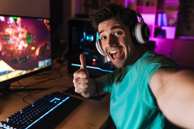 Neşeli bir adamın evde bilgisayarda oyun oynarken baş parmağını göstermesi ve selfie çekmesi.