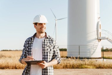 Gülümseyen bir elektrik mühendisi yel değirmenleriyle bir tarlada durup notlar alıyor.