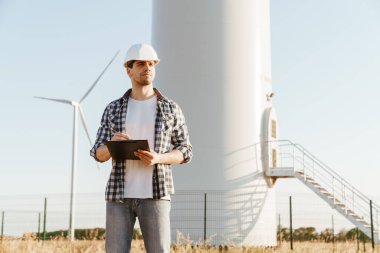 Gülümseyen bir elektrik mühendisi yel değirmenleriyle bir tarlada durup notlar alıyor.