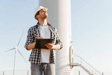 Gülümseyen bir elektrik mühendisi yel değirmenleriyle bir tarlada durup notlar alıyor.