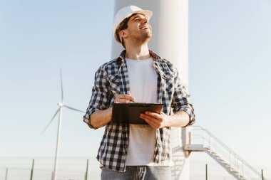 Gülümseyen bir elektrik mühendisi yel değirmenleriyle bir tarlada durup notlar alıyor.