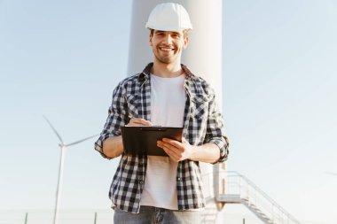 Gülümseyen bir elektrik mühendisi yel değirmenleriyle bir tarlada durup notlar alıyor.