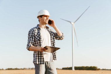 Elektrikli yel değirmeni çiftliğinde dururken cep telefonu kullanan gülümseyen bir erkek mühendis.