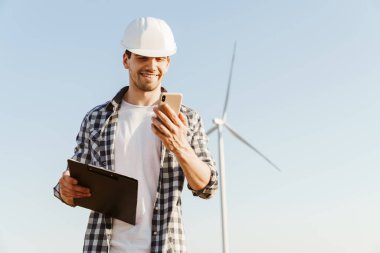 Elektrikli yel değirmeni çiftliğinde dururken cep telefonu kullanan gülümseyen bir erkek mühendis.