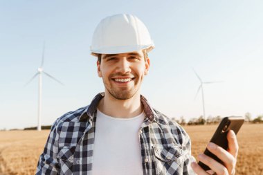 Elektrikli yel değirmeni çiftliğinde dururken cep telefonu kullanan gülümseyen bir erkek mühendis.