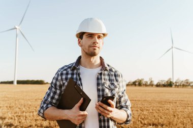 Elektrik yel değirmeni çiftliğinde dururken cep telefonu kullanan bir erkek mühendis.