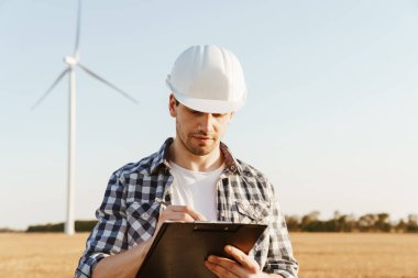 Yel değirmenleriyle dolu bir alanda durup not alan bir elektrik mühendisi.