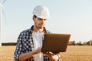 Yel değirmenleriyle dolu bir alanda dizüstü bilgisayar kullanan bir elektrik mühendisi rüzgar türbini enerji üretimini kontrol ediyor.