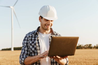 Yel değirmenleriyle dolu bir alanda dizüstü bilgisayar kullanan bir elektrik mühendisi rüzgar türbini enerji üretimini kontrol ediyor.