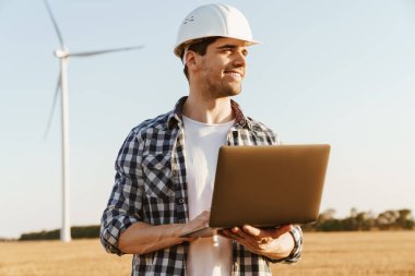 Yel değirmenleriyle dolu bir alanda dizüstü bilgisayar kullanan bir elektrik mühendisi rüzgar türbini enerji üretimini kontrol ediyor.