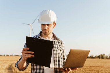 Yel değirmenleriyle bir alanda duran bir elektrik mühendisi dizüstü bilgisayar kullanıyor, pano tutuyor, rüzgar türbini enerji üretimini kontrol ediyor.