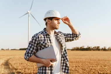 Yel değirmenleriyle dolu bir alanda elinde dizüstü bilgisayarla duran bir elektrik mühendisi rüzgar türbini enerji üretimini kontrol ediyor.