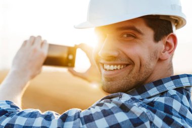 Bir tarlada rüzgar türbinlerinin fotoğrafını çeken kasketli, gülümseyen bir işçiyi yakından çek.