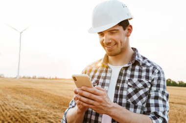 Elektrikli yel değirmeni çiftliğinde dururken cep telefonu kullanan gülümseyen erkek mühendis.