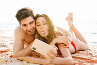 Kumsalda battaniyeye uzanmış selfie çeken mutlu genç çift.