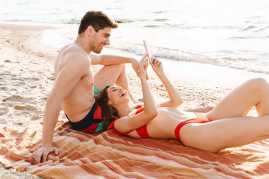 Kumsalda battaniyeye uzanmış selfie çeken mutlu genç çift.