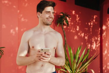 Kumsalda gevşeyen, cep telefonuyla gülümseyen yakışıklı adam.