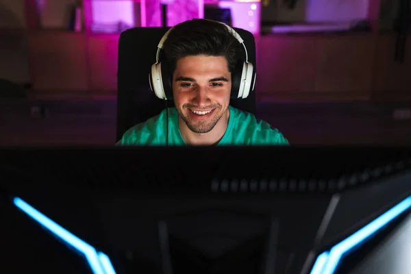 Image of caucasian happy man in headphones playing video game on computer while sitting at table indoors