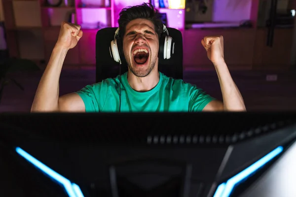 自宅でコンピュータゲームをプレイしながら 勝者のジェスチャーを作るヘッドフォンで興奮した若い男のイメージ — ストック写真