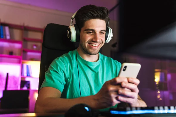Image of caucasian smiling man in headphones using cellphone playing video game on computer at home