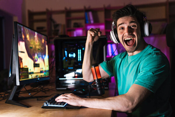 Изображение взволнованного молодого человека в наушниках, делающего жест победителя во время игры на компьютере дома