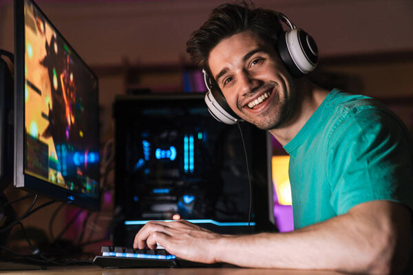 Изображение белого счастливого человека в наушниках, играющего в видеоигру на компьютере, сидящего за столом в помещении