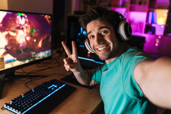 Evdeki bilgisayarda bilgisayar oyunu oynarken barış işareti yapan ve selfie çeken neşeli bir adamın görüntüsü