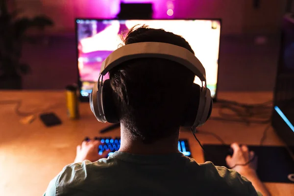 屋内のテーブルに座っている間 コンピュータ上でビデオゲームをプレイヘッドフォンで焦点を当てた男のイメージ — ストック写真