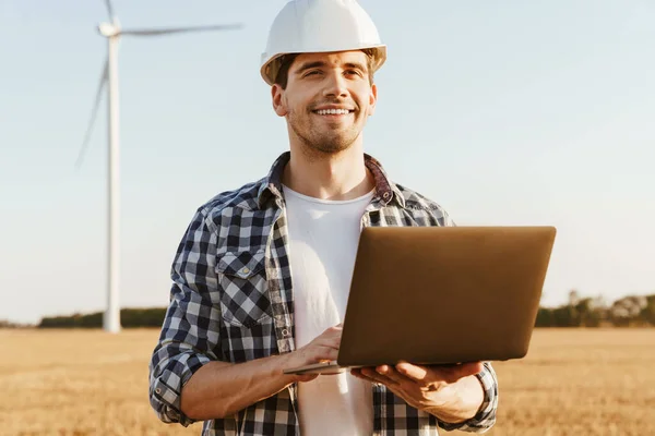An electrical engineer standing on a field with windmills, using laptop computer, checking on wind turbine energy production