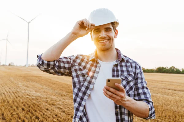Elektrikli yel değirmeni çiftliğinde dururken cep telefonu kullanan gülümseyen erkek mühendis.