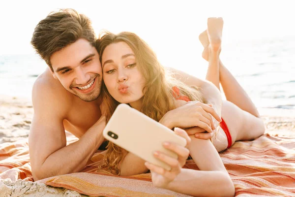 Kumsalda battaniyeye uzanmış selfie çeken mutlu genç çift.