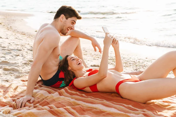 Kumsalda battaniyeye uzanmış selfie çeken mutlu genç çift.