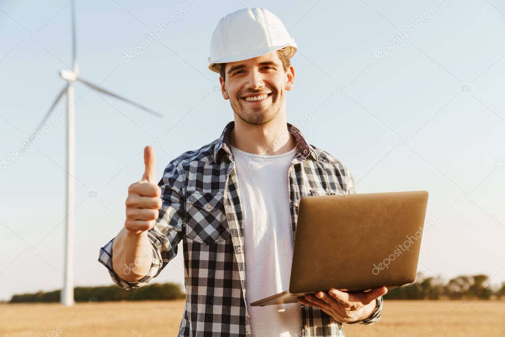 An electrical engineer standing on a field with windmills, using laptop computer, thumbs up