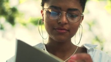 Gözlüklü ve kulaklıklı güzel bir Afro-Amerikan kadının yakın çekim görüntüsü parkta oturan defterinde yazıyor.