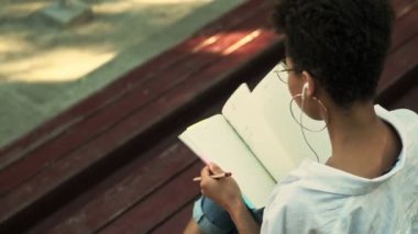Gözlüklü ve kulaklıklı Afro-Amerikalı genç bir kadının arka planı parktaki bankta otururken notlar alıyor.