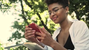 Gözlüklü, gülümseyen Afro-Amerikan bir kadın parktaki bankta oturan akıllı telefonunu kullanıyor.