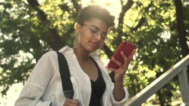 Gülümseyen Afro-Amerikan bir kadın parkta yürürken akıllı telefonunu kullanıyor.