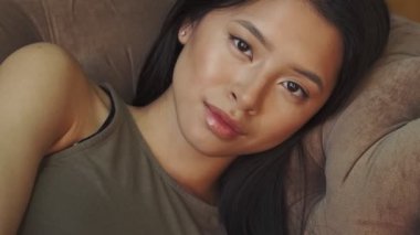 Genç ve güzel bir Asyalı kadın evdeki kanepede uzanmış kameraya bakıyor.