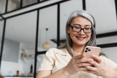 Olgun, neşeli iş kadınının evdeki koltukta otururken cep telefonu kullanmasının fotoğrafı..