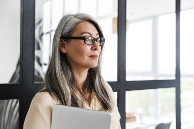 Ciddi, olgun bir iş kadınının elinde dizüstü bilgisayarla evin penceresinin yanında dikilmesi..