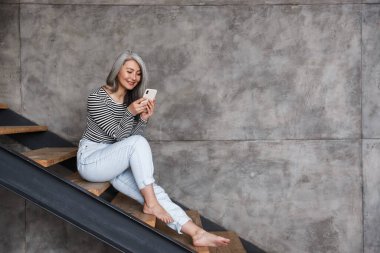 Evde cep telefonu kullanırken merdivenlerde oturan mutlu ve olgun bir kadın görüntüsü.