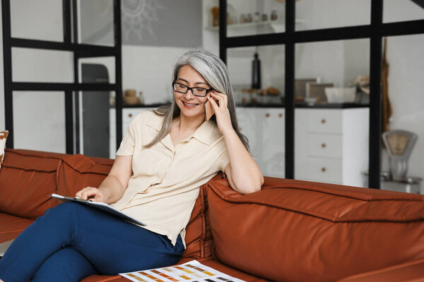 Photo of happy mature business woman sitting on sofa indoors at home while talking by mobile phone.