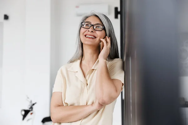リビングで携帯電話で話す眼鏡をかけた白髪のアジア人女性の笑顔の写真 — ストック写真