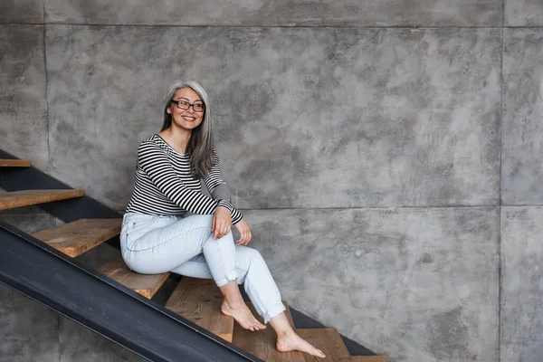照片上 快乐的白发苍苍的亚洲女人坐在家里的楼梯上 戴着眼镜微笑 — 图库照片